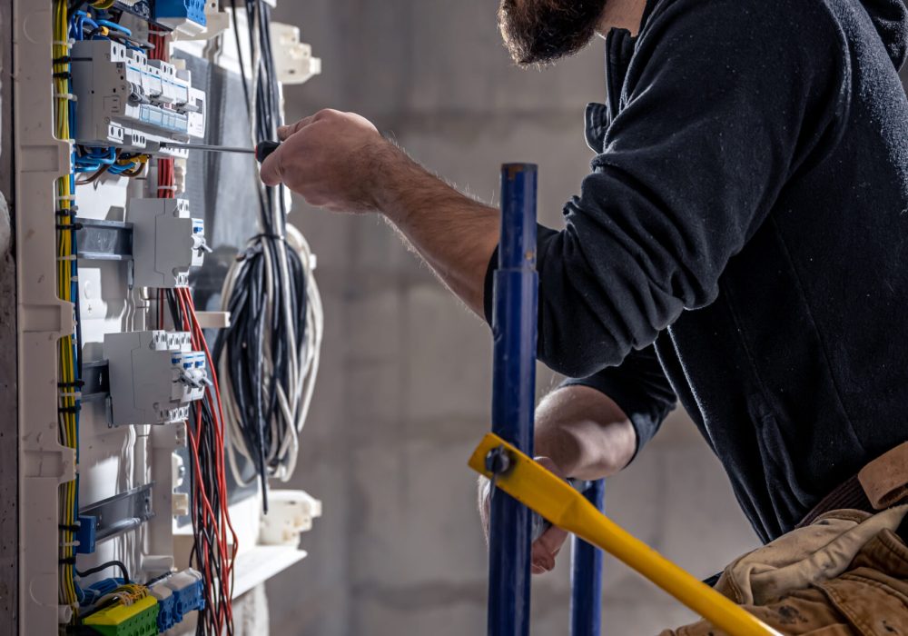 A male electrician works in a switchboard with an electrical connecting cable, connects the equipment with tools.